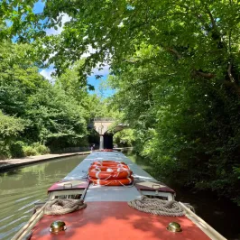 Canal boat with lifebuoys navigating under a leafy bridge.