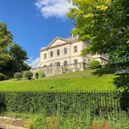 Elegant white house on a hill surrounded by trees and greenery under a blue sky.