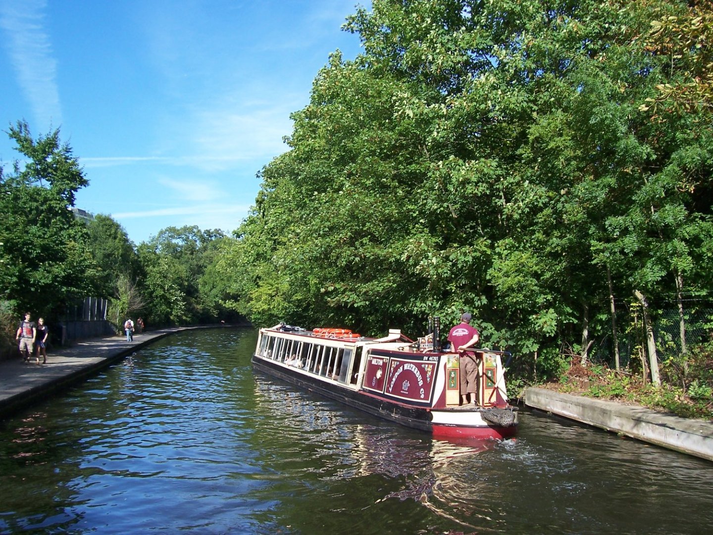 Narrowboat on a tree-lined canal with people walking along the path.