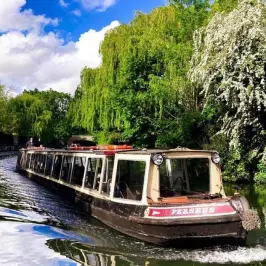 Long narrowboat on a canal, surrounded by green trees under a blue sky.