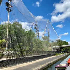 Enclosed netted aviary near a canal with a blue sky and bridge in the background.