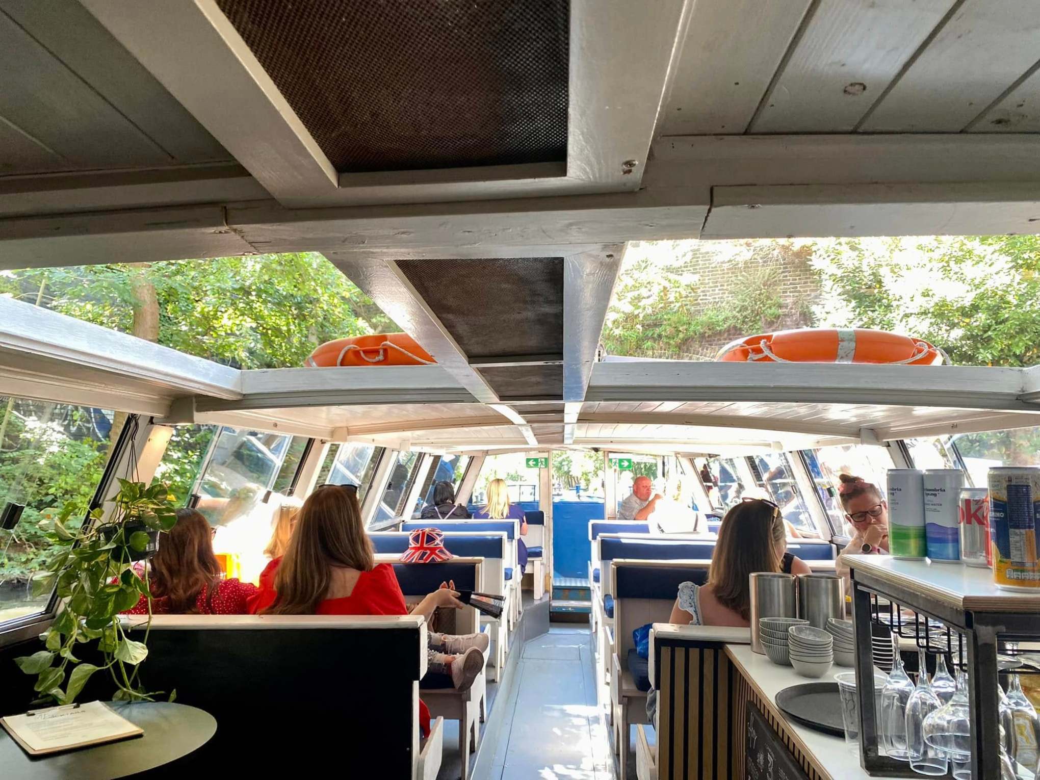 Interior of a boat with people seated at tables, lifebuoys on roof, and trees visible through windows.