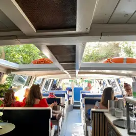 Interior of a boat with people seated at tables, lifebuoys on roof, and trees visible through windows.