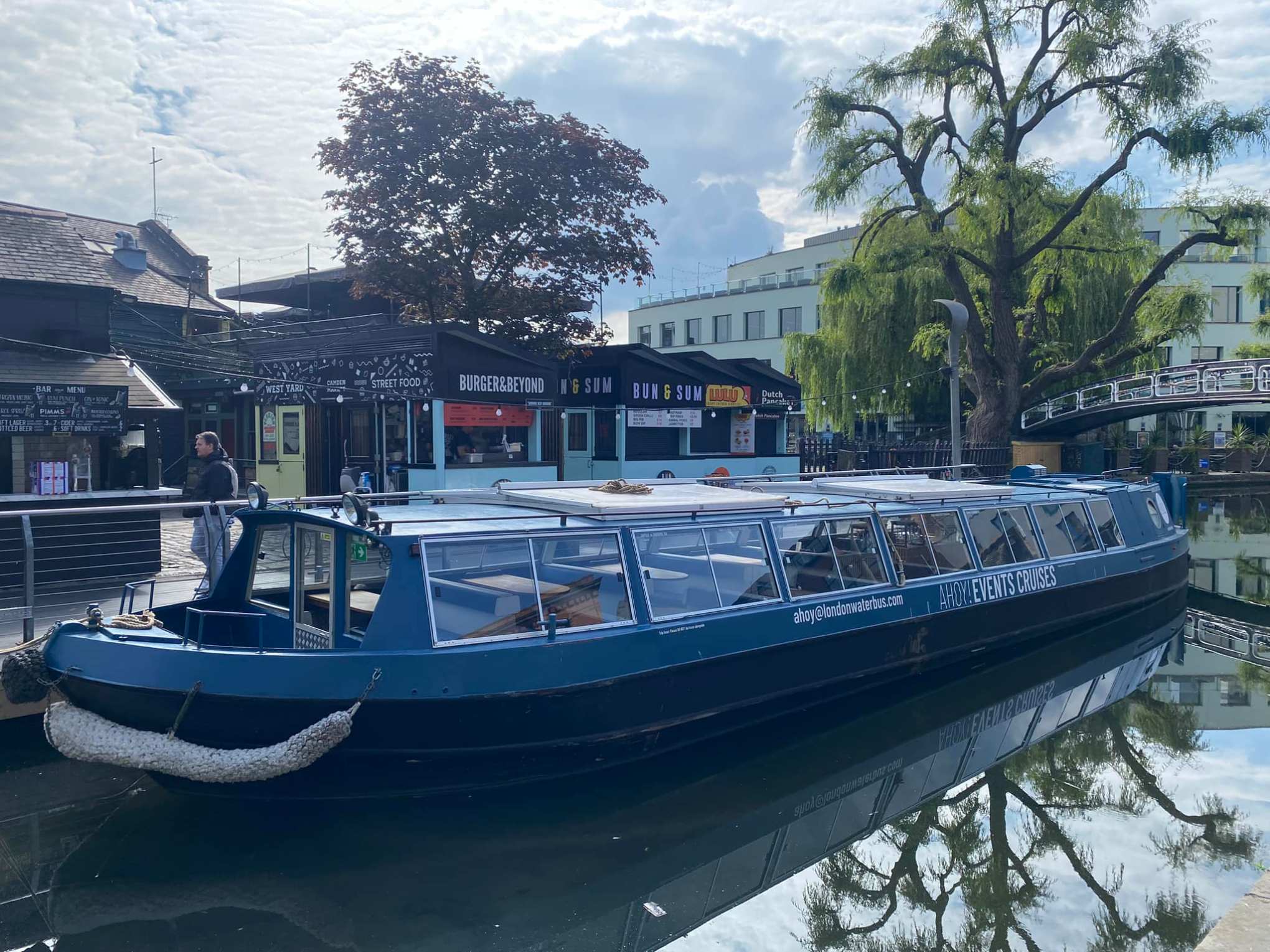 Blue canal boat docked near food stalls and a tree-shaded walkway.