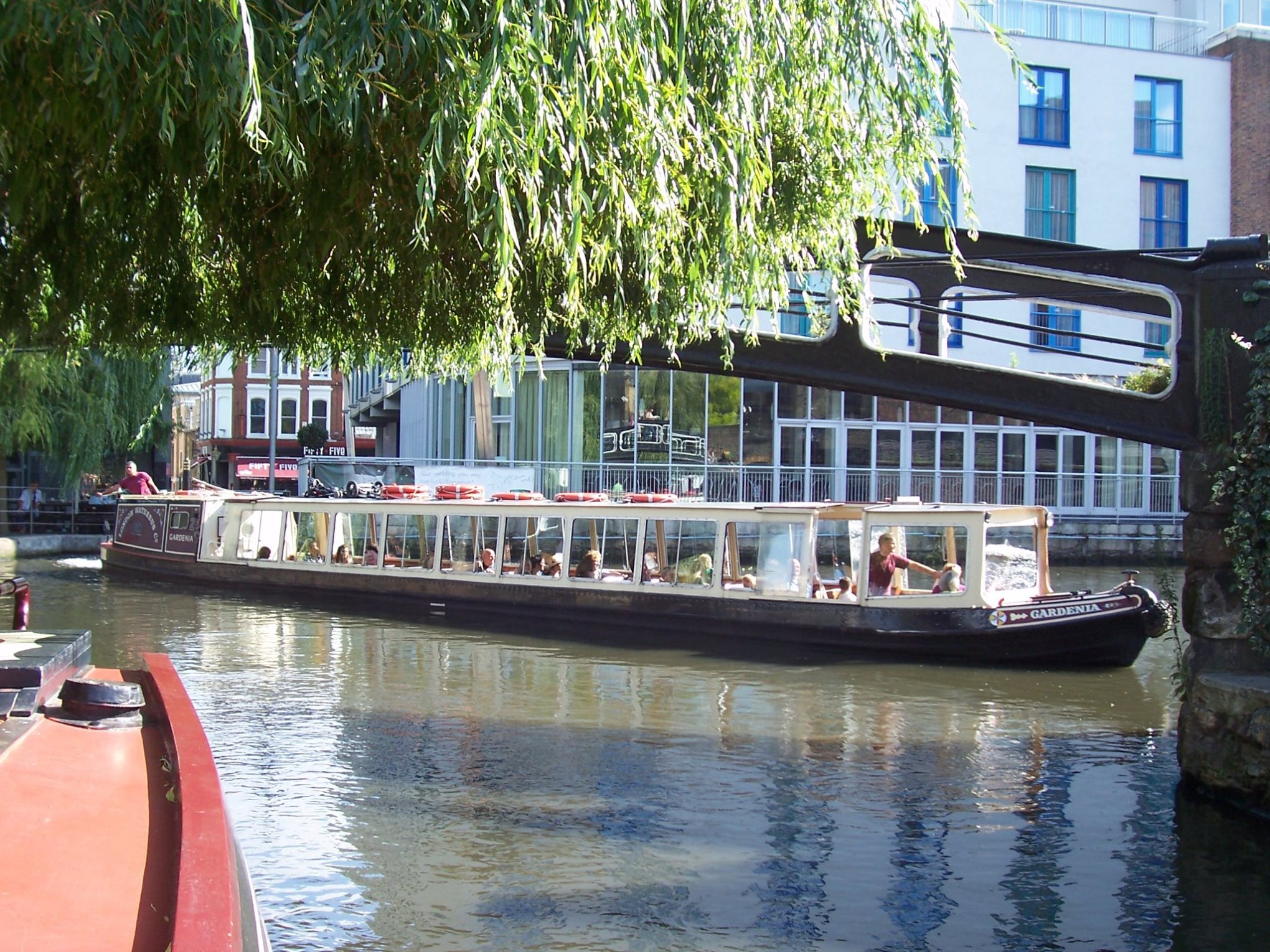 Riverboat with passengers under willow tree near modern buildings on a canal.