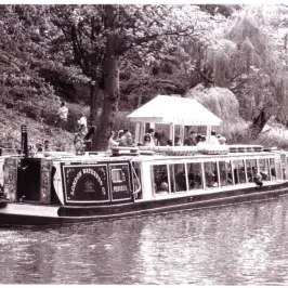 Vintage boat on canal with passengers, surrounded by trees and path along the water.