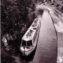 A canal boat travels along a narrow, tree-lined canal near a building and pathways.