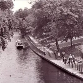 Black and white photo of a tree-lined canal with a boat and people walking along the path.
