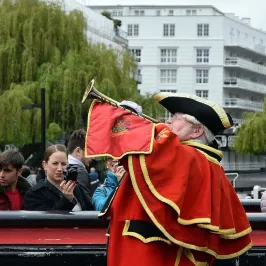 Man in red historical costume playing trumpet, with onlookers photographing outdoors.