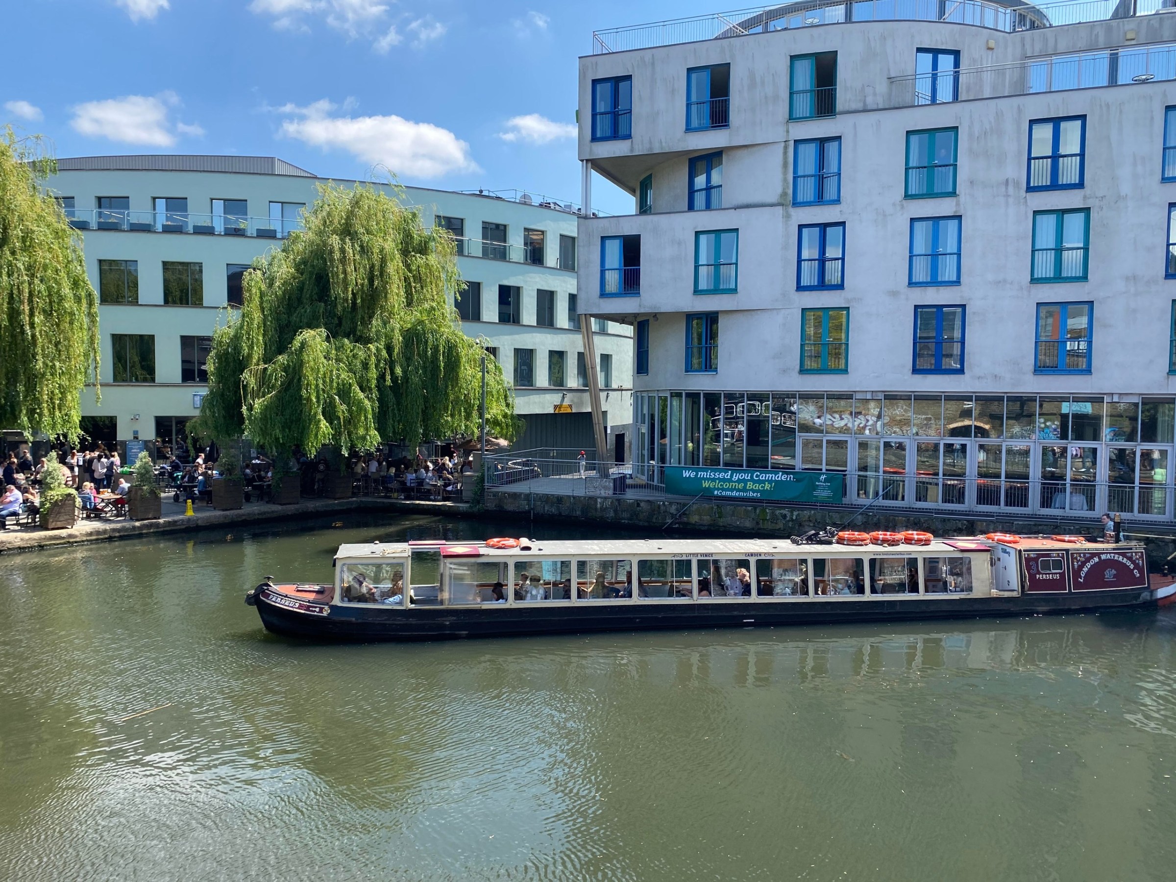 Canal boat on a river with modern buildings and lush trees under a blue sky.