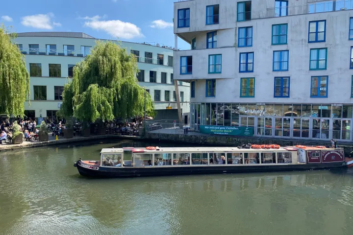 Canal boat on a river with modern buildings and lush trees under a blue sky.