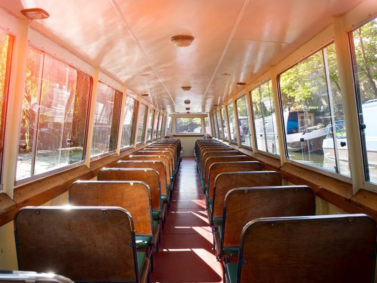 Interior of an empty canal boat with rows of wooden seats and large windows.