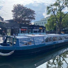 Blue canal boat moored near street food stalls and a tree, with a bridge in the background.