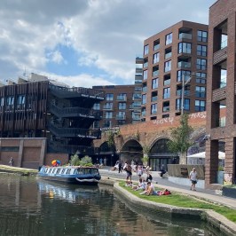 Urban canal scene with modern brick buildings, people walking, and a narrowboat on the water.