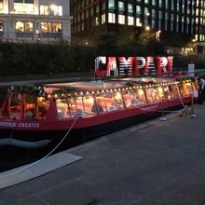 Red boat named 'CA'CREATES' moored at night, decorated with lights, with city buildings in the background.