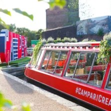 Red boat with plants and #CampariCreates moored next to geometric art structure on canal.
