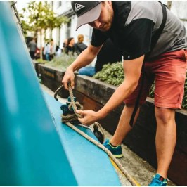Man tying a rope on a boat next to shrubs, wearing a cap, t-shirt, and shorts.