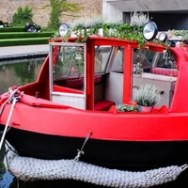 Red boat with plants on deck moored on a canal, green steps in background.