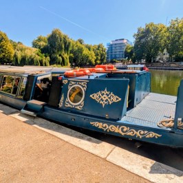 A blue canal boat docked on a sunny riverside with trees and buildings in the background.