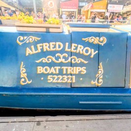 Blue boat with decorative text: 'Alfred Leroy Boat Trips 522321' docked at a busy market.